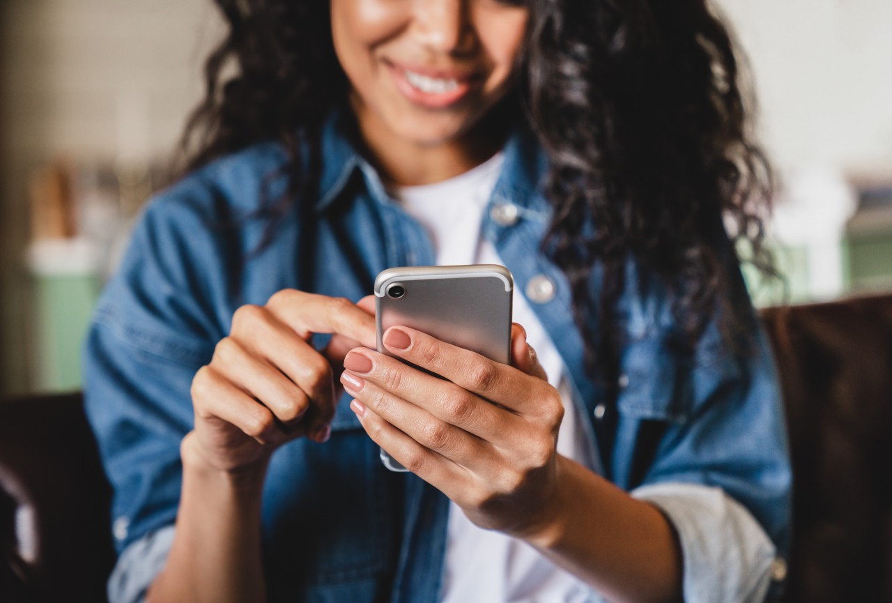 A woman in a denim jacket uses a smartphone, smiling and engaging with the device.
